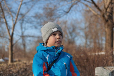 A boy stands in a park wearing a bright blue jacket and looks away on a sunny day in winter or early...