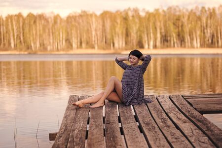 young cute girl sits on a pier in a summer dressの写真素材