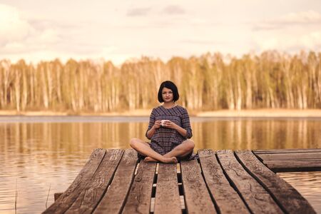 young cute girl sits on a pier in a summer dressの写真素材