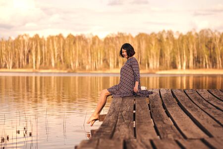young cute girl sits on a pier in a summer dressの写真素材