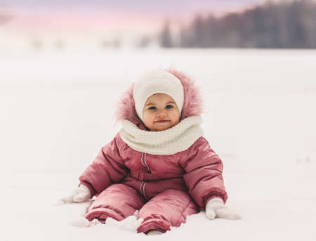 A cute little girl sits in the snow in the winter outdoorsの写真素材