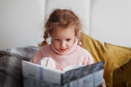 Beautiful little girl reads a book sitting on the couchの写真素材