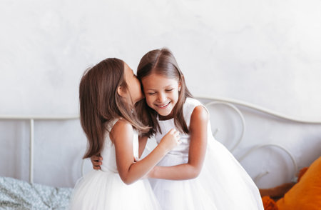 A little girl whispers in her sister's ear. Two girls girlfriends in a light room