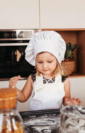 A little cute girl in a white chef hat plays with flour on the kitchen tableの写真素材