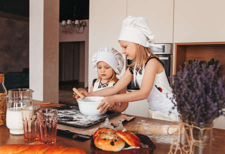 Two little girls make dough in the kitchen together. Sisters play in white hats of the chief