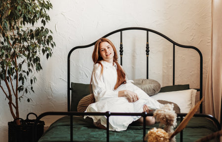 Young happy smiling girl with long hair sits with a pillow on the bed in the bedroom