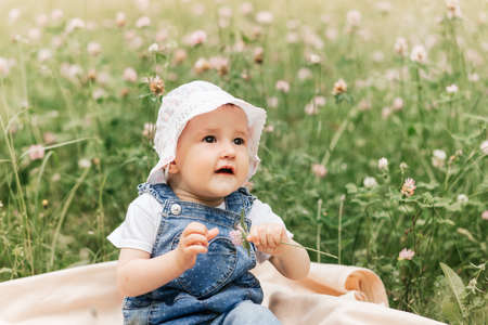A little infante girl in a white cap sits in the park among the flowersの写真素材