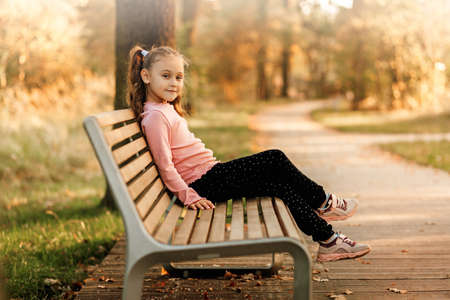 Little pretty girl sits on a bench in the park in the autumnの写真素材