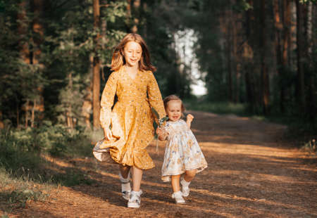Two sisters run through the forest holding hands. Summer sunny day and girls in light dresses.