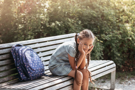 Back to school. A cute little schoolgirl in a dress with pigtails and large blue backpackis sitting on a bench in the school yard. A little girl is going to the first grade.の写真素材