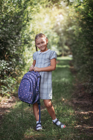 Back to school. A cute little schoolgirl in a dress with pigtails and large blue backpackis walks in the school yard. A little girl is going to the first grade.の写真素材