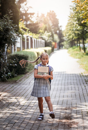 Back to school. A cute little schoolgirl in a dress with pigtails and large blue backpackis holds books and stand in the school yard. A little girl is going to the first grade.の写真素材
