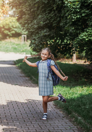 A little girl in a dress with pigtails holds large blue backpack and walks on road alley. First september day in elementary school.の写真素材