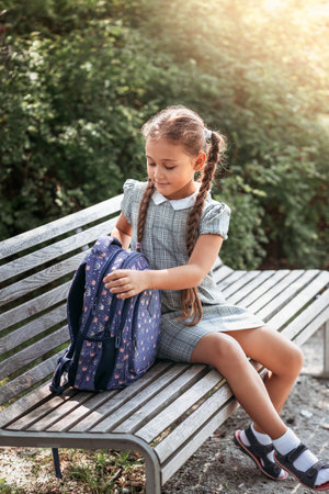 Back to school. A cute little schoolgirl in a dress with pigtails and large blue backpackis sitting on a bench in the school yard. A little girl is going to the first grade.の写真素材