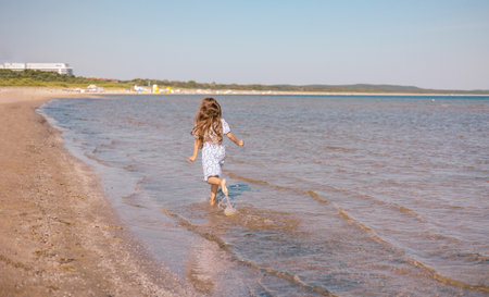 Little girl with lond hair in beautiful dress running on sea beach during summer holiday.Child playing on ocean beach. Sea vacation for family with kids.の写真素材