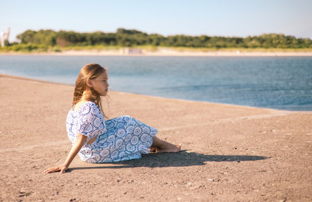 little girl in a dress on the beach with developing long blond hair in the wind and looking on the seaの写真素材