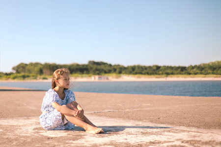 little girl in a dress on the beach with developing long blond hair in the wind and looking on the seaの写真素材