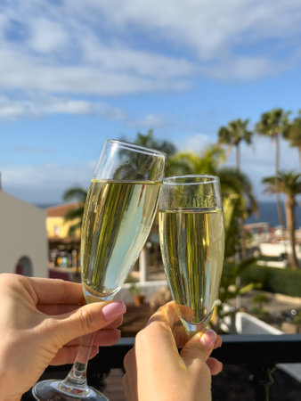Two glasses of champagne in women's hands against the backdrop of a tropical resort. A toast to a successful vacation and the beginning of the holidaysの写真素材