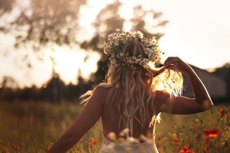 Young blonde woman in a wreath of daisies on a flower field at sunsetの写真素材