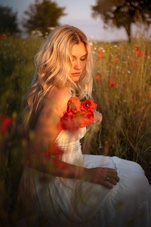 Portrait of a young beautiful blonde woman with red poppy flowers in her hands on a flower field in summerの写真素材