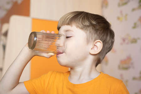 cute little boy drinking water from glass indoors on blurred orange background. Childhood, healthy lifestyle.の写真素材