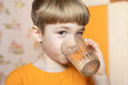 Close up portrait of cute little boy drinking water from glass indoors on blurred background. Childhood, caring for children, healthy lifestyleの写真素材