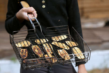 woman hand holds barbecue grill with vegetables fried eggplant on fire in garden