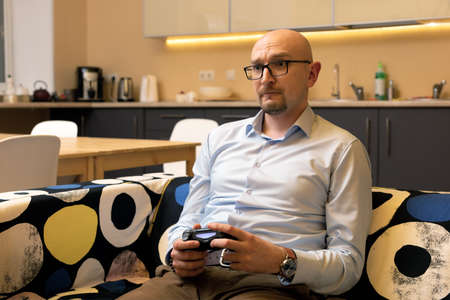 middle-aged bald man with glasses is playing video games with joystick at home. Businessman relieves...