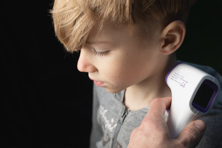 Measuring boy's temperature using non contact device infrared thermometer gun on dark backgroundの写真素材