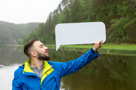 Portrait of happy young caucasian man holding empty banner or message bubble over nature background