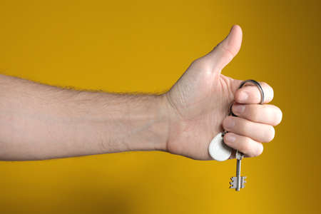 male hand holding house keys and showing thumb up on yellow background. Concept of successful real...