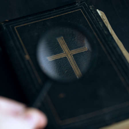 Vintage antique Bible book on table and magnifying glass in human hand. Research historical religious documents with loupe, wisdom word search.の写真素材