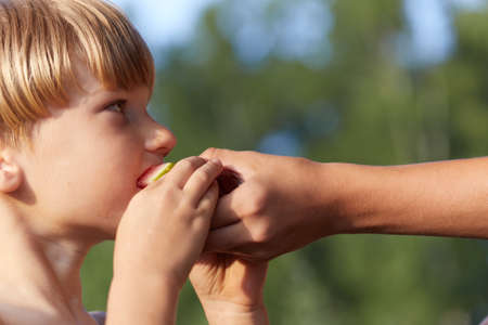 Little boy kid eating watermelon closeup side view. Male child with european face appearance biting red juicy melon in mother or sister hand. Natural summer scene background with blurred effectの写真素材