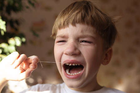 photo of crying 6 years boy who is trying to tear up his milk tooth by thread. Feeling of pain and...