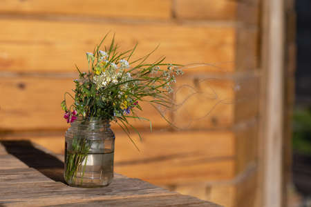 Modest bouquet of white and pink wildflowers with green twigs in glass jar is placed on wooden table near wall of house outside on blurred natural background. Summer still life. Pleasant memories.の写真素材