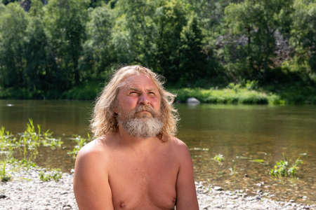 Portrait of naked mature long-haired bearded man carefully looking at sky sitting on shore of on natural background of pond and forest. Looking hopeful. Meditation or prayer in nature.の写真素材