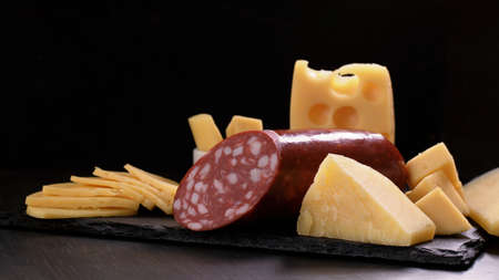 Several types of cheese and piece of salami loaf are on black stone board on dark background....