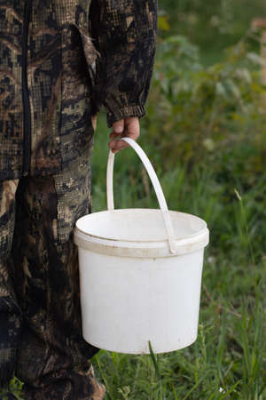 Close-up of white plastic bucket with place for writing in hands of man in camouflage suit on natural green background. Paint or building material. Worker, soldier, hunter or fisherman with bucket.の写真素材
