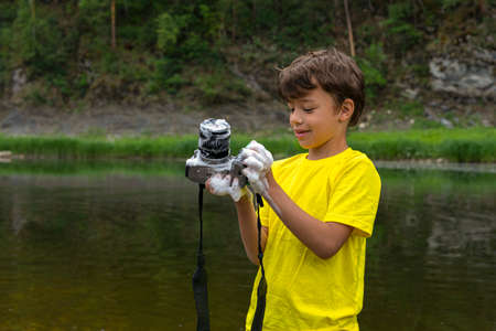 Naughty kid standing on bank of mountain river holding digital camera he had just washed with soap. Half-length portrait, copy space. Childhood, childish prank, while mom was distracted, mischiefの写真素材
