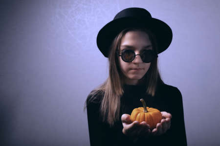 Young girl wearing black hat and black glasses. Small orange pumpkin in her hands. Black clothes add some horror to picture and creepy atmosphereの写真素材