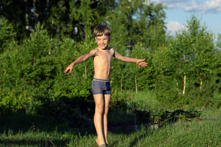 Happy smiling boy in wet shorts plays with water sprinkler on natural blurred background. Summer games on vacation. happy life.の写真素材