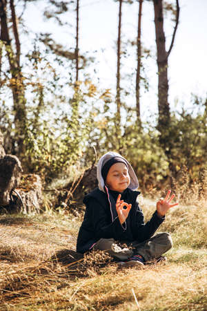 A boy in warm clothes meditates in the forest while sitting on the grass while walking. Portrait of a childの写真素材