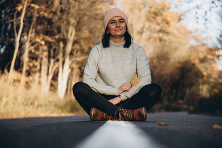 Portrait of a girl who sits on an empty road in the autumn forest. Forest at sunsetの写真素材