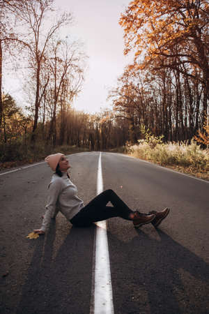 Portrait of a beautiful smiling girl who sits on an empty long road in the autumn forest. Sunset time. Front viewの写真素材