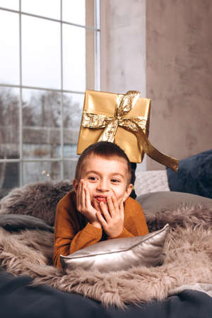 A boy in a yellow sweater is lying on the bed, and behind him is a gift wrapped in gold. Portrait of a boyの写真素材