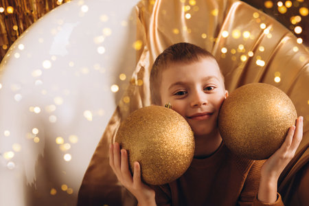 The boy holds two large golden Christmas balls in his hands and smiles. Portrait of a child in Christmas lightsの写真素材