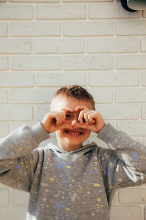 The boy smiles and shows a heart with his fingers. Portrait of a child against a white brick wall. Close upの写真素材
