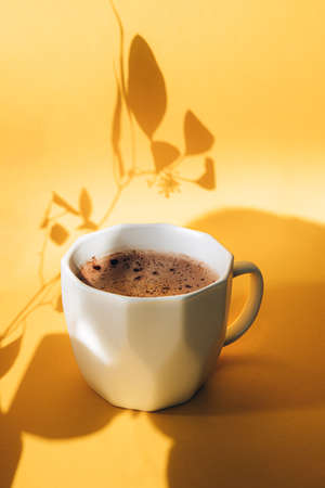 White cup with coffee on a yellow background with shadows. Front view