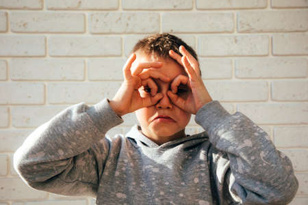 A boy on the background of a brick wall looks through his fingers imitating binoculars with a sad face. Portrait of a schoolboyの写真素材