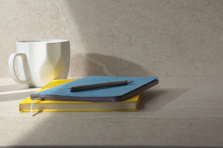 Two notebooks, yellow and blue, and white cup on a beige table with a pen. Office table and...
